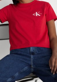 T-shirt rossa in cotone con scollo rotondo, caratterizzata da un logo bianco di Calvin Klein Jeans sul petto. Abbinata a jeans di denim blu scuro.