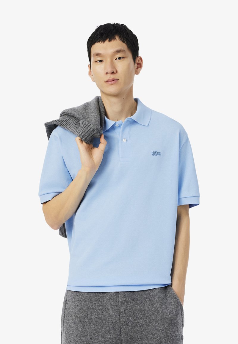 Ljusblå kortärmad polo med krage, med en subtil broderad logotyp. Grå tröja hängande över modellens axel.