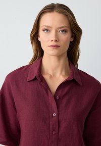 Giovane donna con pelle chiara e occhi verdi che indossa una camicia rossa scura a maniche lunghe, con dei bottoni, rivolta in avanti con un'espressione neutra.