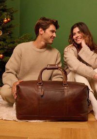 Rjava usnjena športna torba z dvema ročajema, sprednjim žepom z zadrgo in vidnim šivom. Postavljena na mehko belo preprogo z zelenim ozadjem.