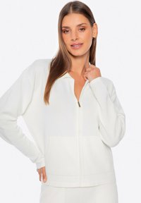 Chaqueta blanca con cremallera y mangas largas, de tela texturizada y con bolsillos frontales. Tiene una pequeña cremallera dorada y presenta un ajuste casual y relajado.