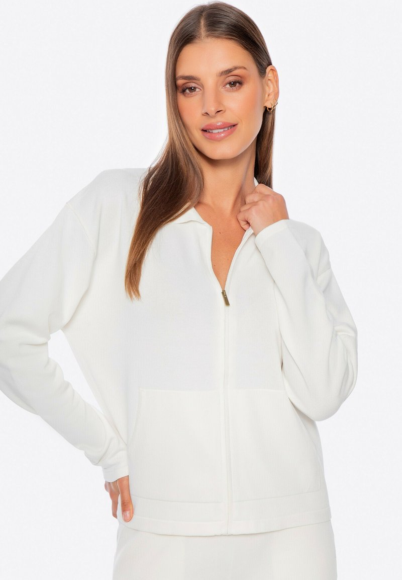 Chaqueta blanca con cremallera y mangas largas, de tela texturizada y con bolsillos frontales. Tiene una pequeña cremallera dorada y presenta un ajuste casual y relajado.
