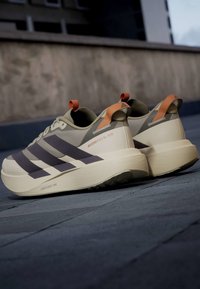 Ζεύγος μπεζ και γκρι παπούτσια Adidas για τρέξιμο με πορτοκαλί ταινίες έλξης πάνω σε πεζοδρόμιο από πέτρα δίπλα σε τσιμεντένιο τοίχο.
