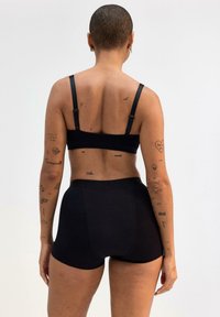 Bralette in morbido tessuto nero e pantaloni corti a vita alta, con spalline regolabili. Tono della pelle visibile con vari tatuaggi sulle braccia.