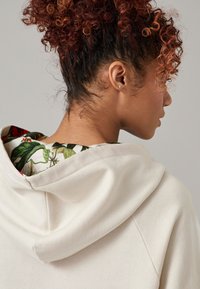 Beige Kapuzenpullover mit verstellbarem Kapuzenzug, ausgestattet mit einem floral gemusterten Innenfutter in Grüntönen und Rot. Weicher Stoff mit lässiger Passform.