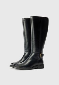 Des bottes en cuir noirs jusqu'au genou présentent une finition lisse, un bout rond, de subtils surpiqûres sur le côté et une boucle décorative sur la cheville extérieure.