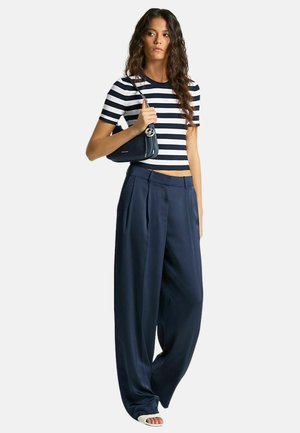 Donna che indossa un top a maniche corte a righe nere e bianche e pantaloni a gamba larga blu navy, che tiene una piccola borsa blu navy con tracolla a catena.
