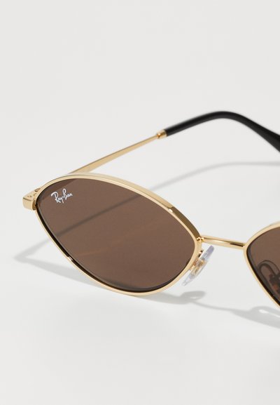 Lunettes de soleil en métal doré avec des verres marron, de forme ovale, avec des plaquettes de nez transparentes et des embouts de branches noirs. Logo présent sur le verre.