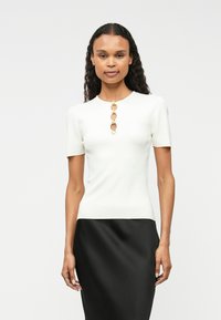 Biała, prążkowana bluzka z krótkim rękawem z okrągłym dekoltem i złotymi okrągłymi guzikami ozdobnymi wzdłuż środka. Nosi się ją z czarną spódnicą.