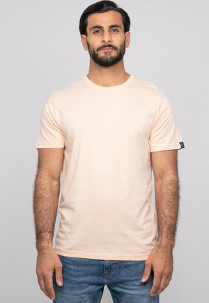 Homme aux cheveux foncés et à la barbe, portant un t-shirt à manches courtes beige clair uni et un jean bleu, debout devant un fond neutre.