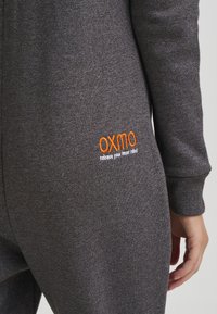 Šedý fleecový overal s oranžovým vyšitým logem značky a textem na zadní straně. Hladké manžety a jemná textura s hladkým povrchem.