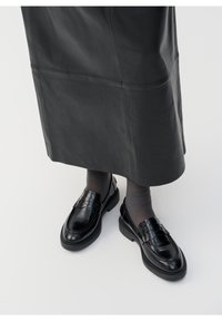 Mocassins en cuir noir avec une finition lisse, présentant une lanière décorative et des coutures contrastantes, associés à des collants gris et une jupe en cuir noir.