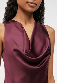 Top de cetim burgundy com decote drapeado; textura suave e design sem mangas; detalhes de hardware mínimos.