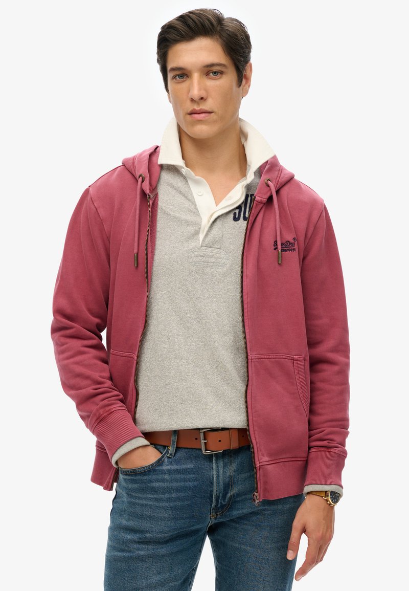 Joven vestido con una sudadera rosa con cremallera sobre una camisa polo gris, jeans azules y un cinturón marrón, de pie con una mano en el bolsillo.