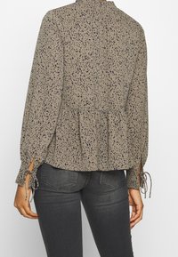 Blusa marrone con motivo a pois blu scuro, vita arricciata, maniche lunghe con lacci e collo alto. Il materiale appare leggero e testurizzato.