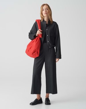 Giacca nera texture con chiusura a bottoni, pantaloni larghi coordinati e mocassini neri; tiene una grande borsa di tessuto rossa.
