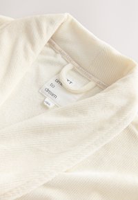 Szlafrok w kolorze kremowym o prążkowanej fakturze, z kołnierzem typu szalowego, wewnętrzną metką marki oraz pętelką do zawieszania z tyłu.