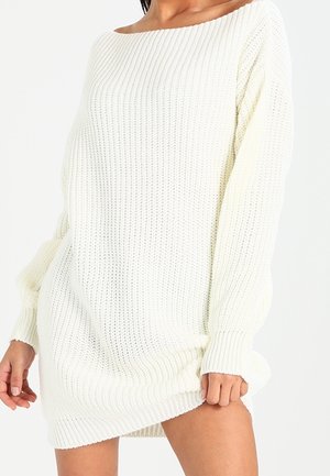 Femme portant une robe-pull en tricot blanc ample à manches longues, tenant l'ourlet et se tenant devant un fond uni.