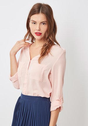 Mujer con cabello castaño ondulado que lleva una blusa rosa claro con mangas enrolladas y una falda plisada azul marino, posando frente a un fondo blanco liso.