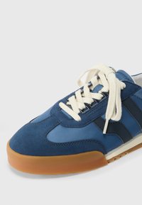 Modrá sportovní bota s designem z nubukové kůže a syntetiky, krémovými tkaničkami, gumovou podrážkou a námořnickými pruhy po stranách.