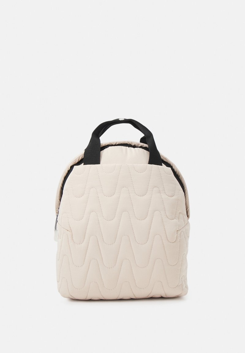 Sac à dos beige matelassé avec poignées noires, un motif de couture ondulé, et une fermeture éclair partiellement visible sur le côté, sur fond blanc.