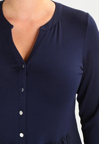 Blouse bleu marine avec un col rond, une fermeture à boutons sur le devant et des manches longues. Confectionnée dans un tissu lisse et léger, avec des détails froncés à la taille.