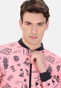 Roze bomberjack met zwarte grafische prints van strandscènes, ananassen en tekst. Geribbelde marineblauwe kraag en ritssluiting.