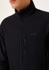 Black softshell jacket with full zip, high collar, and zippered pockets. Features a subtle logo and smooth, stretchy texture.