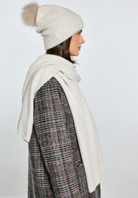 Kremowa czapka z pomponem z futra, w połączeniu z jasnym neutralnym szalem, noszona na teksturowanym płaszczu w kratę w odcieniach szarości i czerwieni.