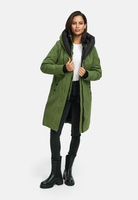 Groene lange parka met een zwarte capuchon, ritssluiting en knoopsluiting, zijzakken en een gladde textuur. Draagt over een paar zwarte jeans en laarzen.