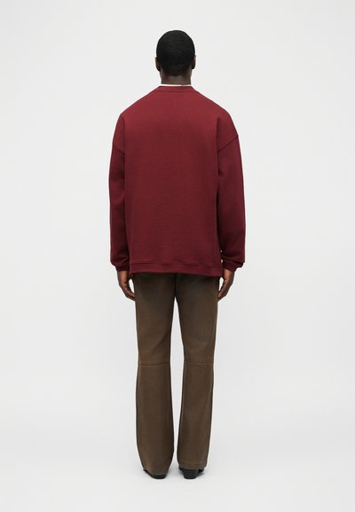 Άτομο που στέκεται με την πλάτη προς το θεατή, φορώντας μπορντό μακρυμάνικη μπλούζα και καφέ παντελόνι ίσιας γραμμής, μπροστά σε απλό φόντο.