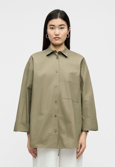 Γυναίκα με ίσια σκουρόχρωμα μαλλιά φοράει φαρδιά πουκάμισο σε ελιά πράσινη απόχρωση με τσέπη στο στήθος και άσπρο παντελόνι μπροστά σε απλό φόντο.