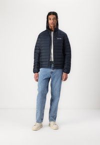 Mørkeblå puffjakke med horisontalt quiltet design, frontlynlås og broderet logo på brystet, kombineret med lyseblå jeans og cremefarvede sneakers.
