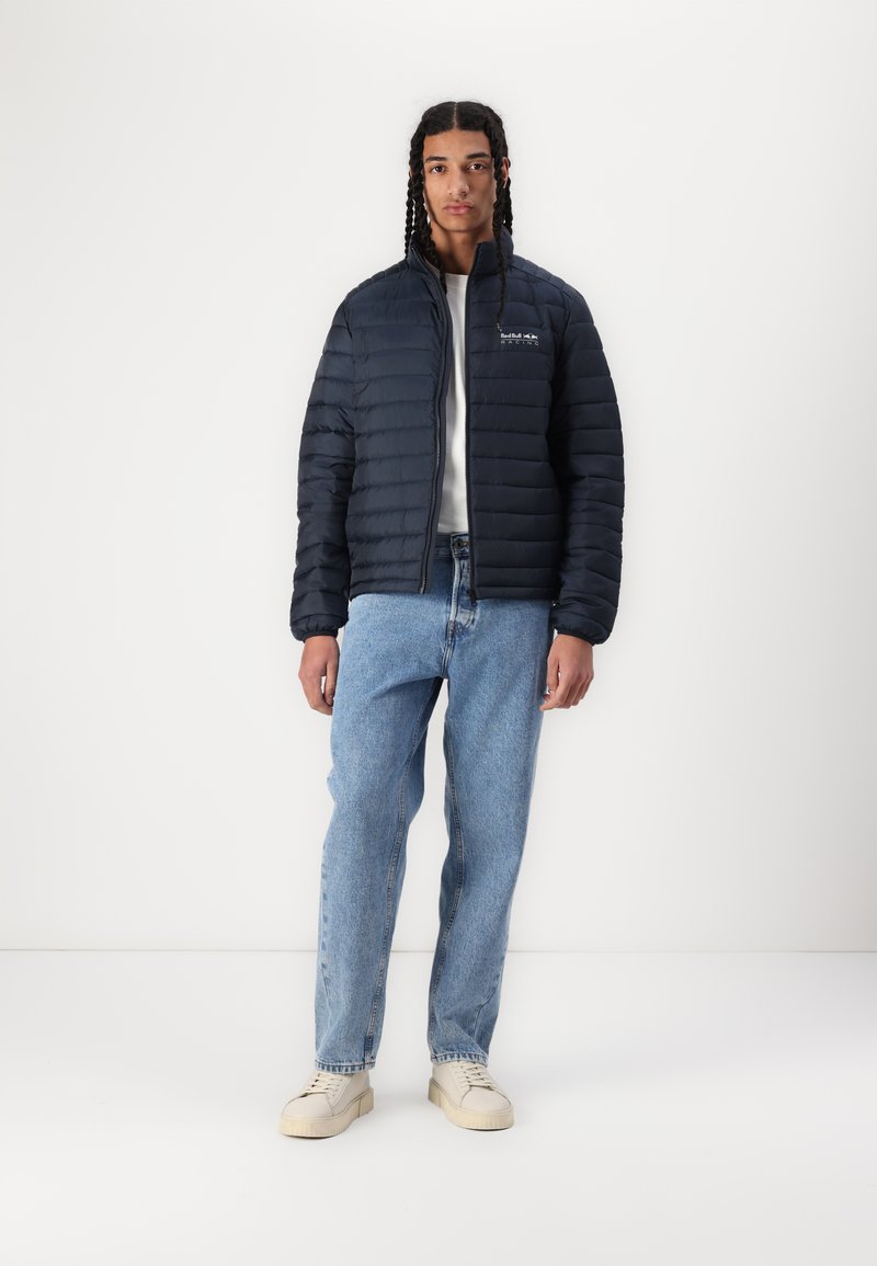 Donkerblauwe pufferjack met horizontaal gequilt ontwerp, rits aan de voorkant en geborduurd logo op de borst, gecombineerd met lichtblauwe jeans en crème sneakers.