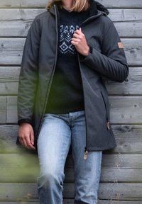 Personne portant une veste à capuche grise sur un pull noir à motifs et un jean bleu clair, debout contre un mur en planches de bois en plein air.