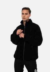 Junger Mann, der den Reißverschluss einer schwarzen Fleecejacke anpasst, vor einem schlichten weißen Hintergrund steht.