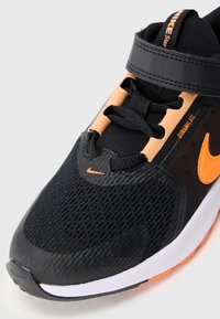 Černá sportovní bota s texturovanou síťovinou na svrchní části, oranžovými akcenty, suchým zipem a bílou podrážkou. Vyznačuje se dynamickým střihem.