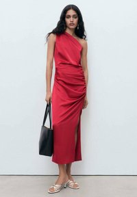 Red, satin, one-shoulder dress with ruching detail and side slit. Paired with a black tote bag and white strappy sandals.