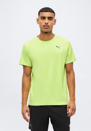Mężczyzna w limonkowym t-shircie marki Puma i czarnych szortach stoi na jednolitym białym tle.
