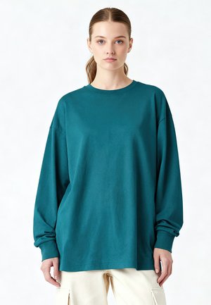 Junge Frau mit zurückgebundenen Haaren, die ein türkisfarbenes, übergroßes Langarmshirt und helle Cargohosen vor schlichtem Hintergrund trägt.