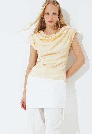 Moteris su ilgais šviesiais plaukais, dėvinti be rankovių šviesiai geltoną tekstūruotą palaidinę, baltą sijoną ir baltas kelnes, stovinti prieš balto fono foną.