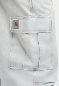 Τσέπη σε ανοιχτό γκρι παντελόνι εργασίας με καπάκι και ραμμένη ετικέτα λογότυπου Carhartt στην πάνω αριστερή γωνία του καπακιού.