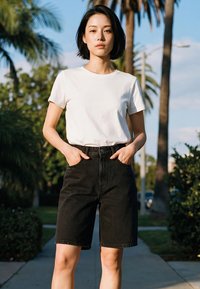 Fiatal nő áll kint egy járdán, kezét fekete farmer rövidnadrágja zsebében, fehér pólót visel, mögötte pálmafák és zöld növényzet látható.