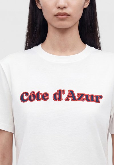 Femme aux longs cheveux foncés portant un t-shirt blanc avec « Côte d'Azur » brodé en rouge et bleu sur la poitrine.
