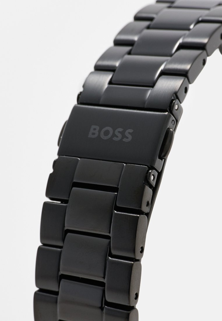 Bracelet de montre en métal noir avec des maillons rectangulaires. Le fermoir présente le nom de la marque "BOSS" gravé sur une surface lisse.