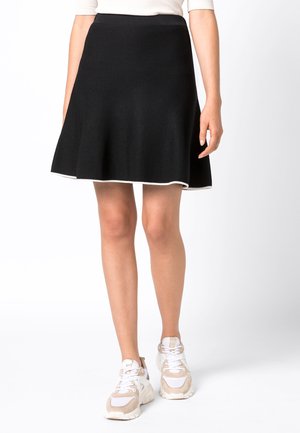 Kobieta ubrana w czarną spódnicę w linii A z białym obszyciem, w połączeniu z beżowo-białymi tenisówkami, stojąca na jednolitym tle.
