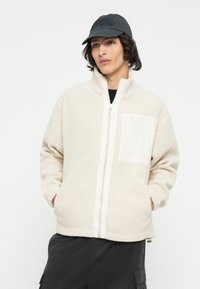 Veste en polaire blanche cassée avec un col haut, fermeture éclair complète et une poche à rabat. Texture douce ; associée à une casquette noire.