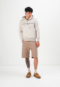 Jongeman in een beige Tommy Hilfiger hoodie, tan shorts, beige sokken en bruine schoenen, staand tegen een effen witte achtergrond.