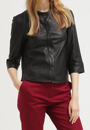 Kobieta ubrana w czarną skórzaną kurtkę o rękawach do połowy przedramienia oraz czerwone spodnie z wysokim stanem, stojąca z jedną ręką w kieszeni.