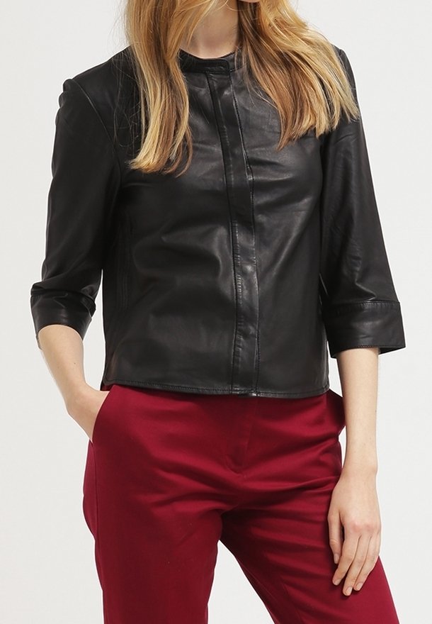 Femme portant une veste en cuir noire à manches trois-quarts et un pantalon rouge taille haute, debout avec une main dans la poche.